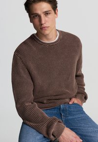 Maglione a maglia marrone con un motivo testurizzato, polsini a coste e scollo rotondo, abbinato a un paio di jeans in denim blu.