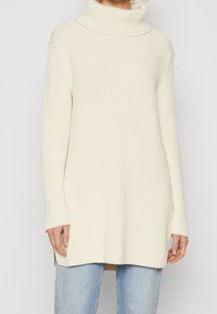 Eine Frau in einem cremefarbenen, gerippten Rollkragenpullover, kombiniert mit hellblauen Jeans, steht vor einem einfarbigen Hintergrund.