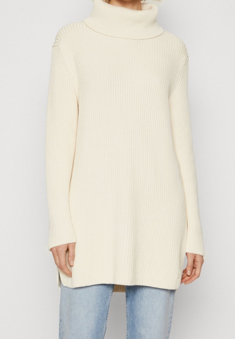 Eine Frau in einem cremefarbenen, gerippten Rollkragenpullover, kombiniert mit hellblauen Jeans, steht vor einem einfarbigen Hintergrund.