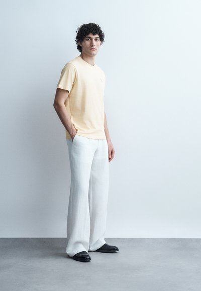 Νεαρός άνδρας με σγουρά μαλλιά στέκεται με τα χέρια στις τσέπες, φορώντας ανοιχτόκίτρινο T-shirt, φαρδιά άσπρα παντελόνια και μαύρα παπούτσια μπροστά σε απλό φόντο.