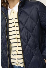 Donna che indossa una giacca trapuntata blu navy su un top a righe crema e blu navy in maglia, con bottoni dorati e un colletto bianco.