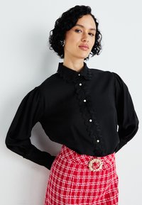 Blusa negra con cuello de volantes y tapeta delantera con botones de perlas, combinada con una falda de cuadros rojos y un cinturón adornado.