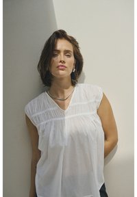 Blouse blanche sans manches avec décolleté froncé et coupe décontractée, présentant une texture subtile. Le mannequin porte un collier en argent sur un fond neutre.