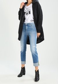Chaqueta negra acolchada con capucha, camiseta blanca con un gráfico de una cámara, vaqueros claros desgastados con puños doblados y botas negras de tobillo.
