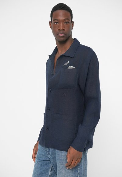 Mlad moški nosi mornarsko modro srajco s platiškom in svetle modre kavbojke.