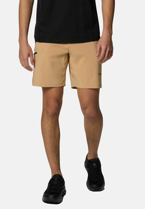 Beige Shorts aus leichtem Stoff, mit seitlichen Taschen und einem Logodetail, kombiniert mit schwarzen Sportschuhen und einem schwarzen Shirt.