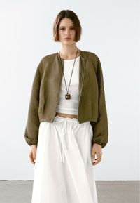 Giubbotto bomber verde oliva con chiusura a zip e polsini elasticizzati, abbinato a un crop top bianco e a una gonna bianca con coulisse. Design minimale.