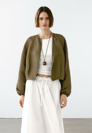 Olivgrüne Bomberjacke mit Reißverschluss und elastischen Bündchen, kombiniert mit einem weißen Crop-Top und einem weißen Rock mit Kordelzug. Minimalistisches Design.