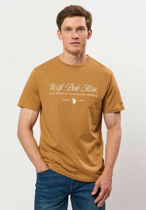 Mann mit braunem U.S. Polo Assn. T-Shirt und blauer Jeans, der mit einer Hand in der Tasche vor einfarbigem Hintergrund steht.