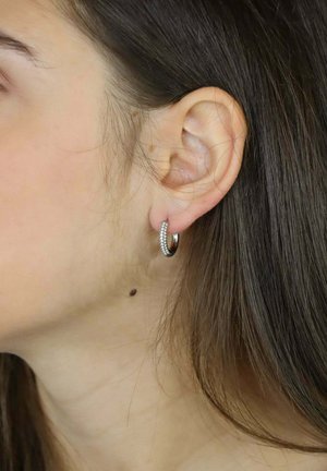 Srebrna kolczyk typu koło z małymi, wtopionymi przezroczystymi kamieniami szlachetnymi wzdłuż przedniego krawędzi, noszony na uchu modela, prezentujący gładką i polerowaną fakturę.