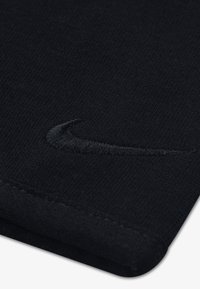 Melnā kokvilnas audums ar strukturētu virsmu un subtilu, izšūtu Nike Swoosh logotipu tajā pašā krāsā, novietotu tuvu malai.