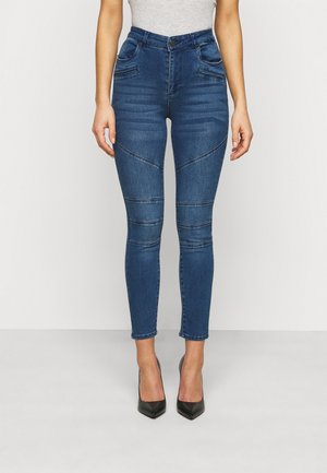 Žena nosící modré džíny s vysokým pasem a úzkými nohavicemi se šitými kolenními panely a černé vysoké podpatky s špičatou špičkou, stojící před jednoduchým pozadím.