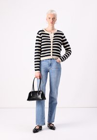 Striped knit cardigan in navy and cream with buttons, paired with light blue jeans. Model holds a small black handbag and wears black flats.