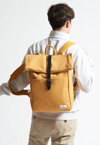 Zaino in tela gialla con design a risvolto, hardware delle cinghie nero e tasca frontale, indossato sulla schiena con spallacci regolabili.
