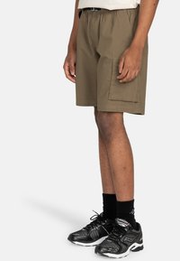 Beige Shorts mit elastischem Bund und seitlicher Cargo-Tasche, locker geschnitten und mit glatter Textur. Kombiniert mit schwarzen Sportschuhen.