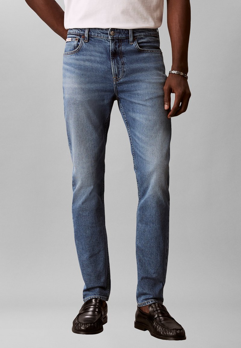 Homme portant un jean slim bleu clair, des mocassins en cuir noir, un t-shirt blanc et un bracelet argenté devant un fond gris uni.