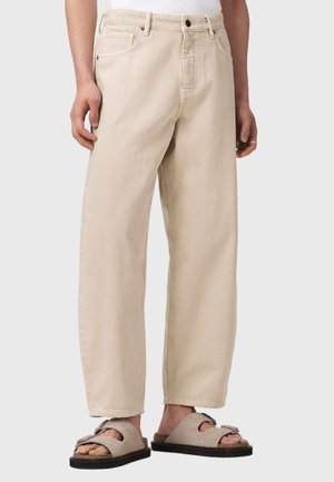 Mann trägt lockere beige Hose und beige offene Sandalen mit zwei Schnallenriemen, steht vor einem neutralen Hintergrund.