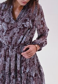 Abito borgogna con motivo paisley, maniche lunghe, vita elastica e tessuto fluido. Accenti argento-blu e dettaglio di orlo arricciato.