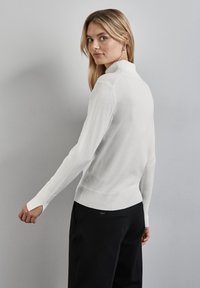 Weißer, figurbetonter Rollkragenpullover mit langen Ärmeln und gerippten Bündchen, kombiniert mit schwarzen Hosen, von hinten vor einem hellgrauen Hintergrund betrachtet.