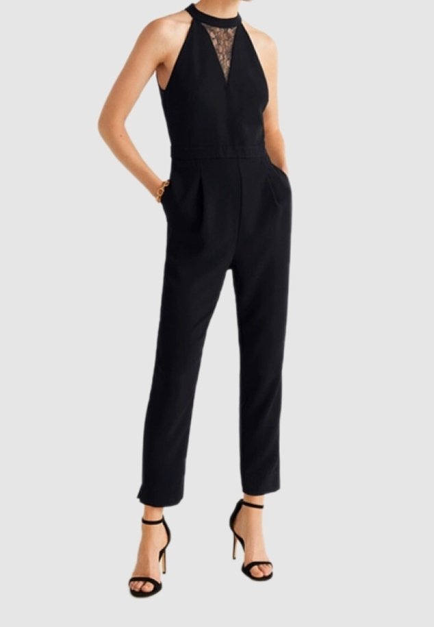 Femme portant une combinaison noire sans manches avec un détail en dentelle au niveau de la poitrine, un pantalon fuselé et des sandales noires à talons hauts, debout les mains dans les poches.