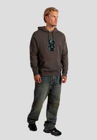 Felpa grigia con cappuccio e tasca frontale, con un design di teschio a cartone animato colorato. Abbinata a jeans blu a vestibilità ampia e scarpe nere.