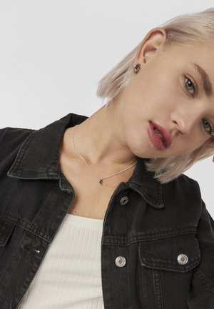 Chaqueta de mezclilla negra con botones plateados, combinada con una blusa blanca de canalé. Collar plateado con un pequeño colgante, cabello rubio corto.