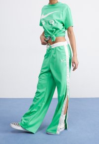 Green Nike crop top with a white logo, paired with green track pants featuring side slits and white stripes. Smooth, shiny fabric.