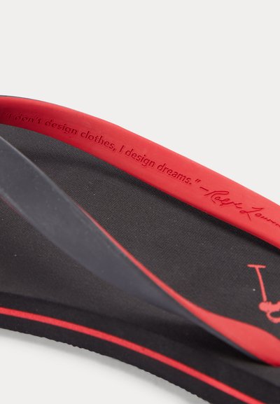 Artimas juodos ir raudonos spalvos šlepetės, kurioje ant dirželio yra iškilus užrašas „Aš nedizainuoju drabužių, aš dizainuoju svajones“.