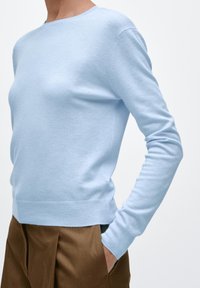 Lichtblauwe trui van zachte stof, met een ronde hals en lange mouwen. Zoom en manchetten zijn geribd. Draagt bijpassende bruine broek.