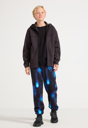 Zwarte hoodie met ritssluiting en twee voorzakken, gecombineerd met een zwarte broek met een patroon van blauwe kwallen. Model draagt zwarte sneakers.