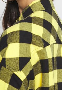 Woman wearing large hoop earring and yellow and black checkered flannel shirt, shoulder and neck visible, long dark hair.
