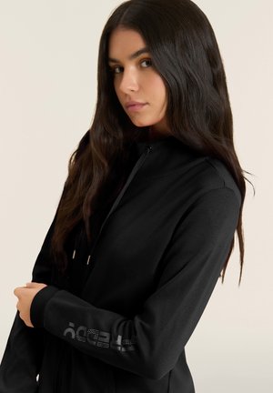 Mujer joven con cabello largo y oscuro, vistiendo una sudadera con capucha negra con cremallera y texto con logo en la manga, posando frente a un fondo liso y claro.