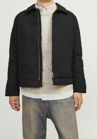 Giacca nera con zip e colletto, indossata sopra un maglione grigio chiaro e una camicia a righe bianche, abbinata a jeans blu.