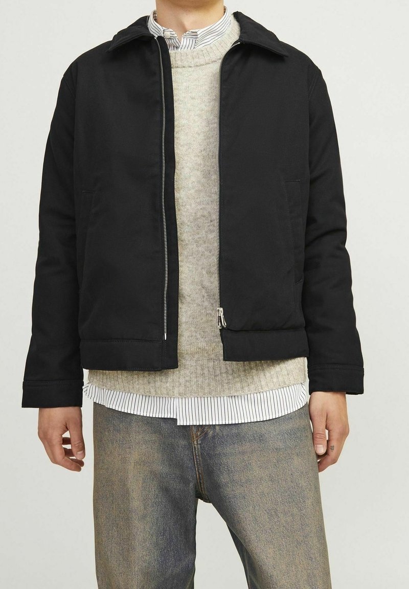Giacca nera con zip e colletto, indossata sopra un maglione grigio chiaro e una camicia a righe bianche, abbinata a jeans blu.
