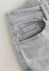 Gros plan sur un jean en denim gris clair montrant la poche avant, la petite poche à monnaie, la passants de ceinture et les rivets métalliques sur un fond clair.