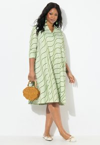 Grünes Kleid mit Wellenmuster, Dreiviertelärmeln und V-Ausschnitt. DieAccessoires umfassen eine geflochtene schalenförmige Tasche und flache weiße Schuhe.
