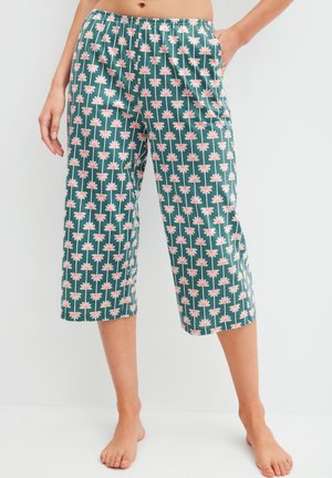 Groene cropped broek met roze en witte bloemmotief, gedragen door een staande persoon met één hand in de zak en blote voeten.
