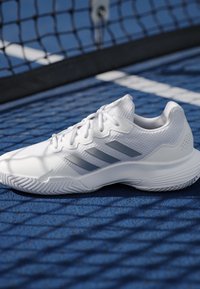 White athletic shoe with gray stripes on blue tennis court near black net and white court lines.