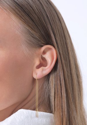 Boucles d'oreilles en chaîne dorée avec un design élégant et allongé, présentant une surface texturée et des éléments suspendus aux côtés de l'oreille d'un modèle.