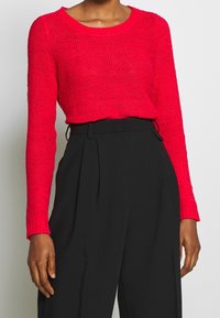 Pull en maille rouge avec un col rond et des manches longues, associé à un pantalon noir taille haute. Le pull présente un motif texturé.