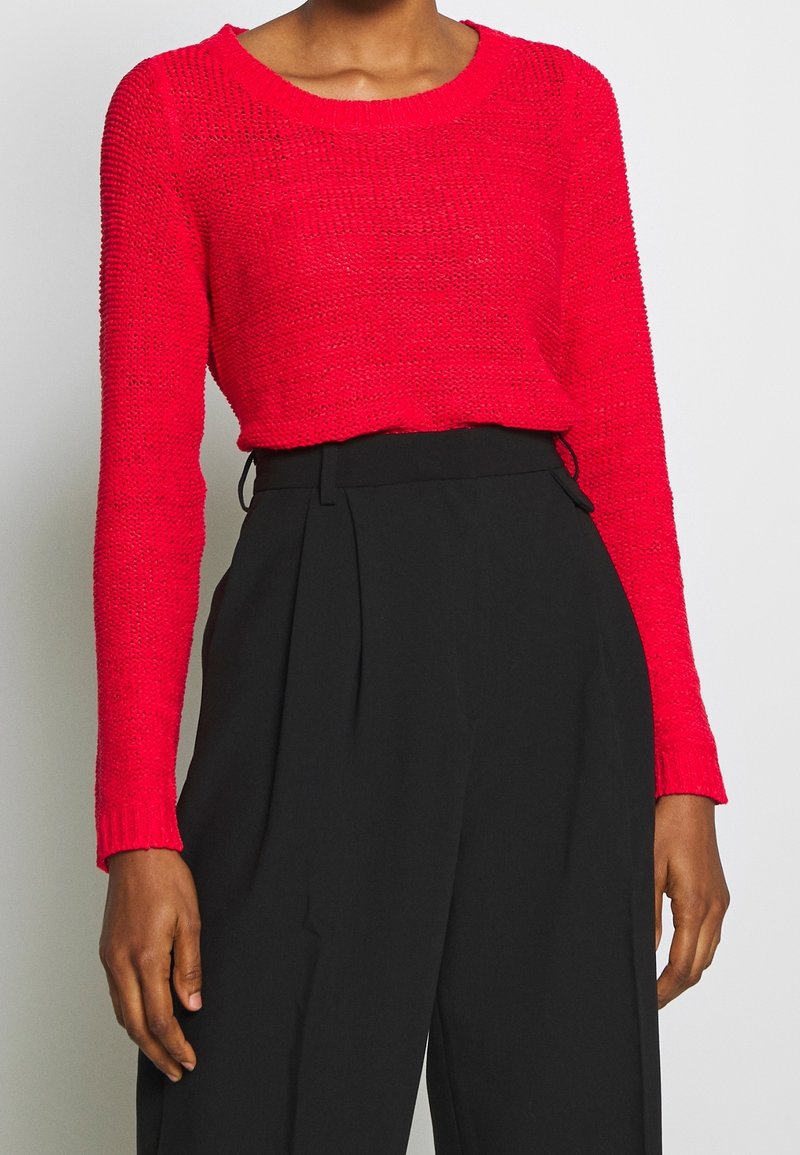 Pull en maille rouge avec un col rond et des manches longues, associé à un pantalon noir taille haute. Le pull présente un motif texturé.