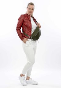 Giacca di pelle rossa con dettagli zip, camicia bianca, borsa a tracolla verde oliva, pantaloni bianchi aderenti e sneakers bianche. Texture lisce e colori a contrasto.