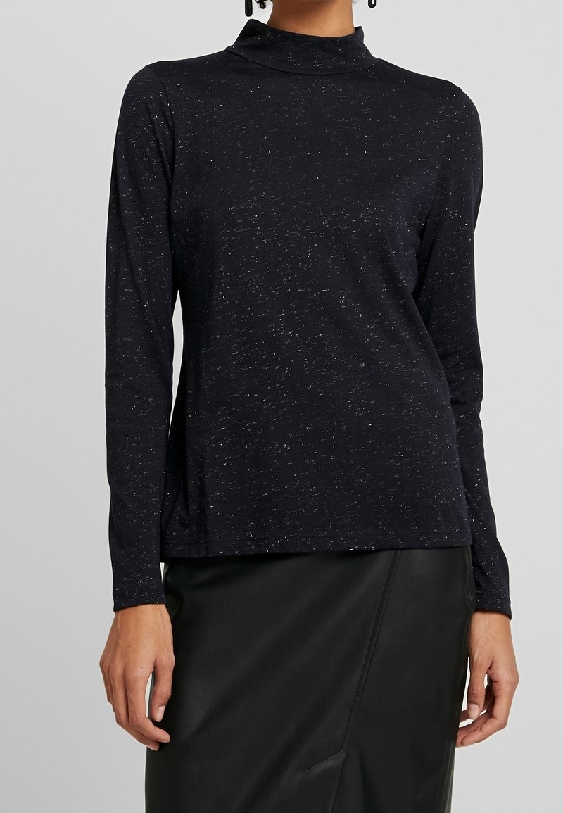Pitkähihautta musta paita, jossa on korkea mock neck, valmistettu teksturoidusta kankaasta, jossa on hienovaraisia metallivihjeitä, yhdistetty mustaan nahkahameeseen.
