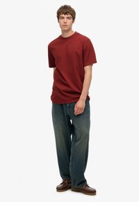 Donkerrode katoenen T-shirt met korte mouwen, ontspannen pasvorm. Gecombineerd met losvallende, verweerde blauwe spijkerbroek en bruine veterschoenen.