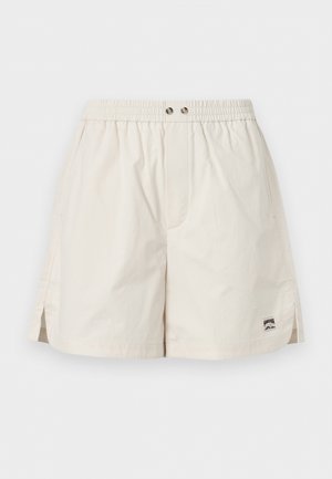 Mladi moški stoji z rokami v žepih, oblečen v modro jakno, karo srajco, bež kratke hlače, kremne nogavice in črne loaferje na enostavni ozadju.