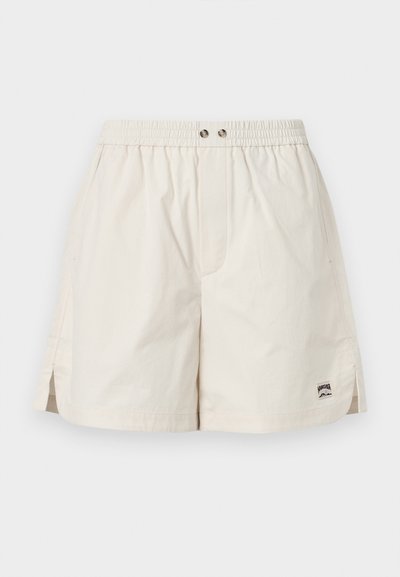 Shorts beige à taille élastique avec fentes latérales, deux boutons métalliques et une petite étiquette de marque rectangulaire sur l'ourlet inférieur droit.