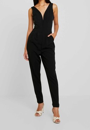 Kobieta ubrana w bezrękawnikowy czarny kombinezon z głębokim dekoltem w serek i zwężanymi nogawkami, w parze z beżowymi sandałami na wysokim obcasie z kokardkami.