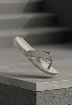 Chancla Nike verde claro con suela texturizada flotando sobre una superficie de piedra gris, con escaleras de concreto desenfocadas al fondo.