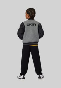 Veste bomber DKNY grise et noire avec des manches bouffantes, pantalon de survêtement noir et baskets blanches, vue de dos.