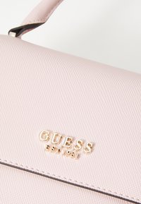 Rausvos spalvos tekstūruota rankinė su auksiniu "GUESS EST 1981" logotipu. Pasižymi sklandžiu, struktūrizuotu dizainu ir siuvinėjimo detalėmis išilgai kraštų.